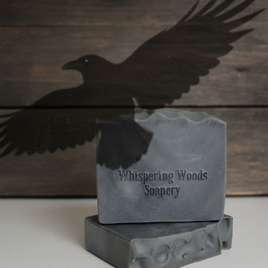 Bars of gray soap with 'Whispering Woods Soapery' branding. There is a shadow of a raven on a wooden background.