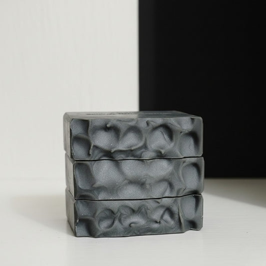 Three gray textured soap bars stacked on a light surface with a black and white background