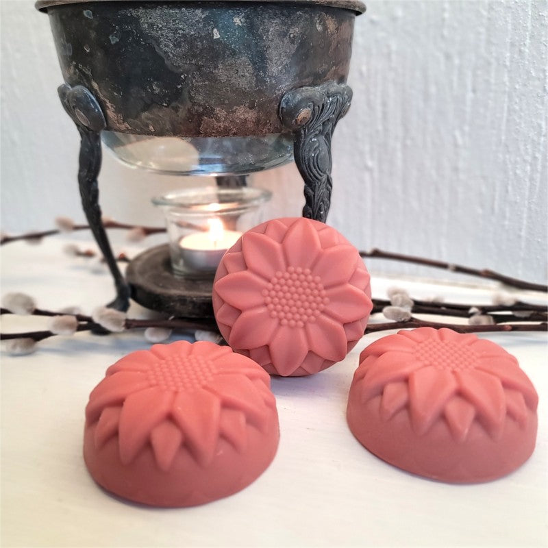 Three dusty-rose-pink flower-shaped soaps arranged in front of a vintage warmer with a lit tealight on a white surface with willow branches.
