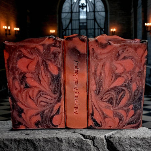 Three soap bars with red and black swirls and 'Whispering Woods Soapery' branding on a stone surface with lit candle sconces on the walls behind them