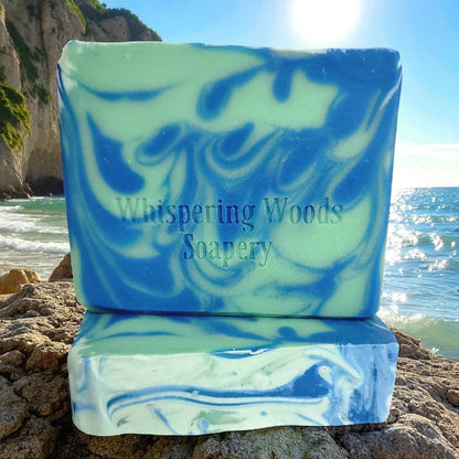 Marbled blue and green soap with 'Whispering Woods Soapery' branding on a rocky beach with water in the background