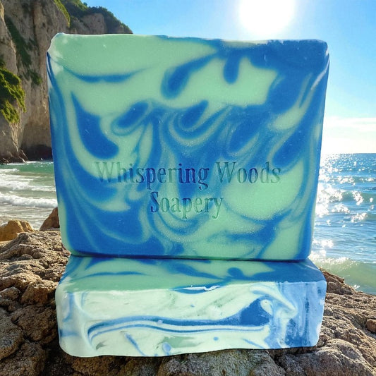 Marbled blue and green soap with 'Whispering Woods Soapery' branding on a rocky beach with water in the background