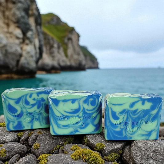 Three bars of 'Whispering Woods Soapery' soap in hues of blue and green sit on rocks with a scenic ocean background