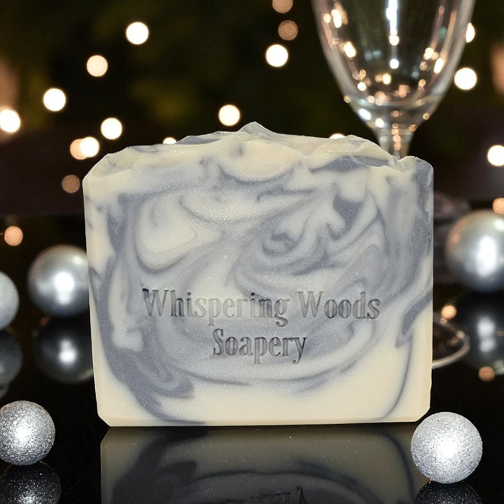 A white bar of soap with silvery swirls with 'Whispering Woods Soapery' branding on a reflective surface with blurred lights in the background