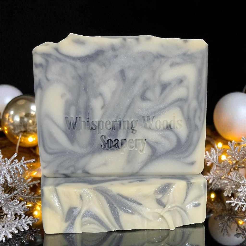 Silver and white soap bar with 'Whispering Woods Soapery' branding on a reflective surface with decorative items in the background