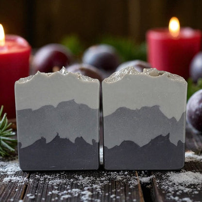Two bars of soap on a wood table with dustings of sugar and a couple lit candles in the background