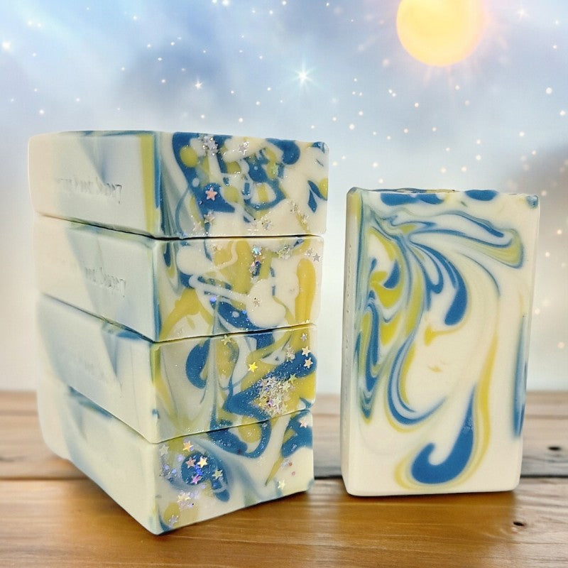 Five white bars of soap with yellow and blue swirls sit on a wood table. The background is a pastel-colored sky with stars and a beaming sun.