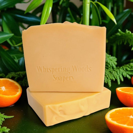 Two bars of light orange soap from 'Whispering Woods Soapery' with tangerines and green foliage in the background