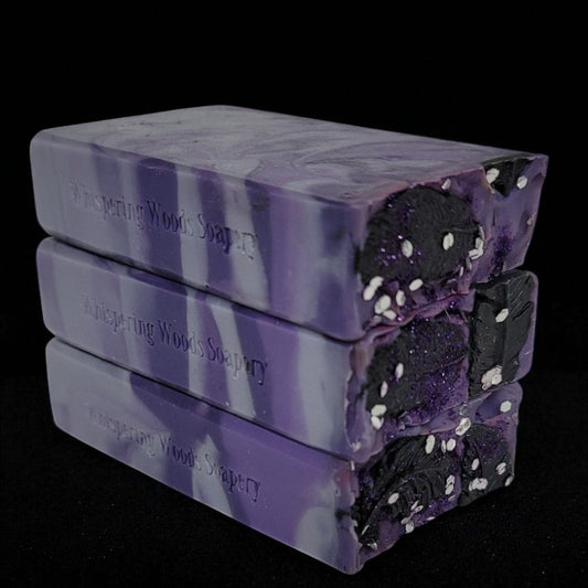 Stack of purple, silver and black soap bars with 'Whispering Woods Soapery' branding on a black background