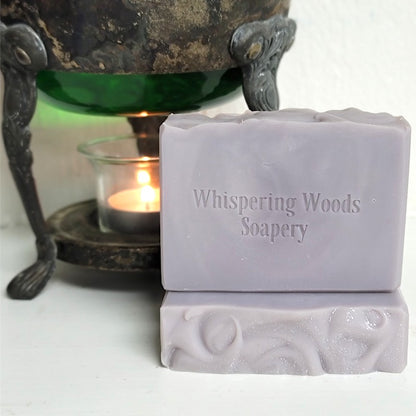 Two silver-purple colored bars of soap stacked on a white surface, with the top bar stamped with the brand name 'Whispering Woods Soapery.' A lit tealight candle in a glass holder sits behind it on a dark metal stand, with a green glass vessel partially visible above.