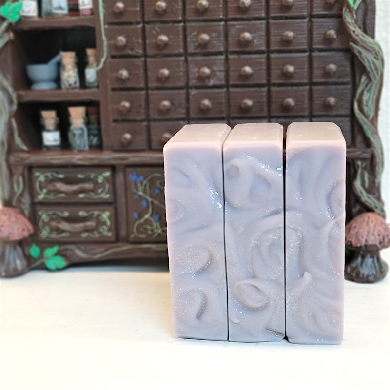 Three rectangular silver-purple soap bars stacked on their sides, showing the sparkle and swirl of their top. They stand in front of a miniature dark brown apothecary cabinet on a white surface.