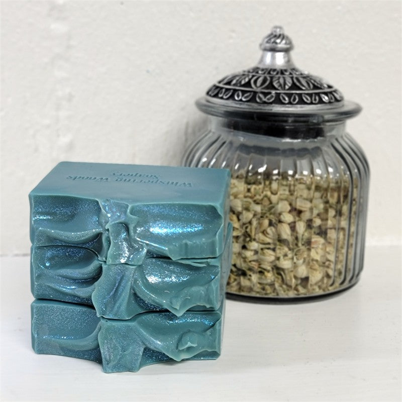 Handmade rectangular bars of teal soap with shimmery sparkle on top stacked on a white surface in front of a clear ribbed glass jar with a silver-toned lid that contains dried botanicals.