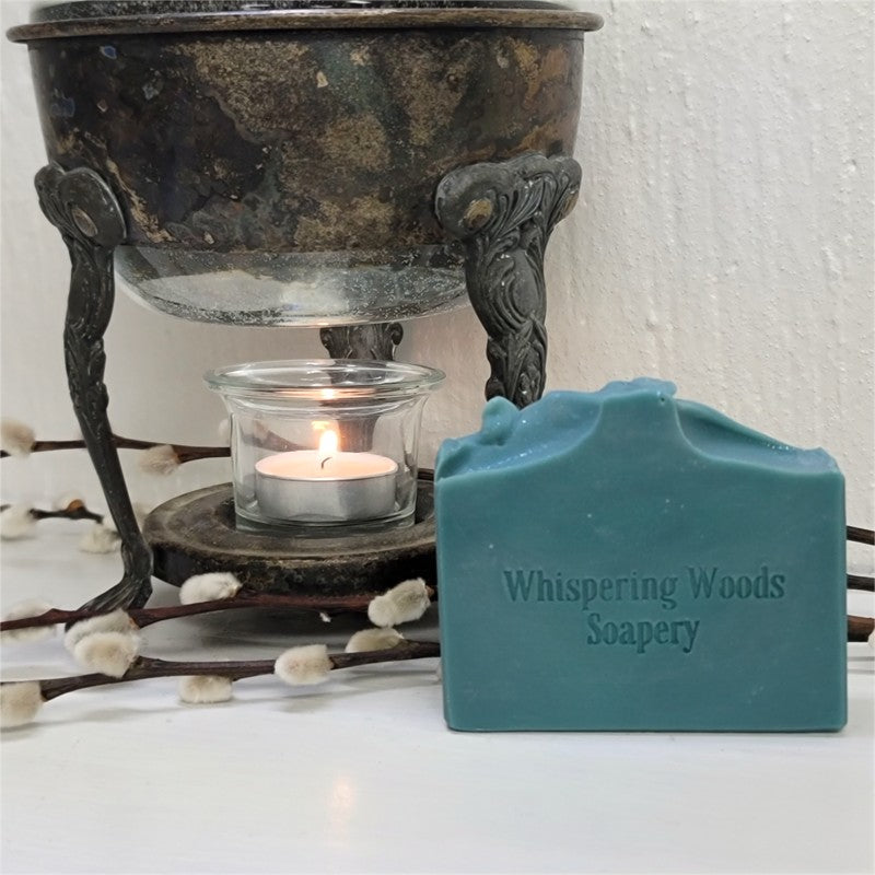 A teal blue bar of soap stamped with 'Whispering Woods Soapery' sits next to a lit glass candle holder on an ornate dark metal burner against a white wall.