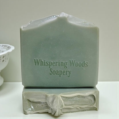 Two bars of soap with 'Whispering Woods Soapery' branding on a white surface