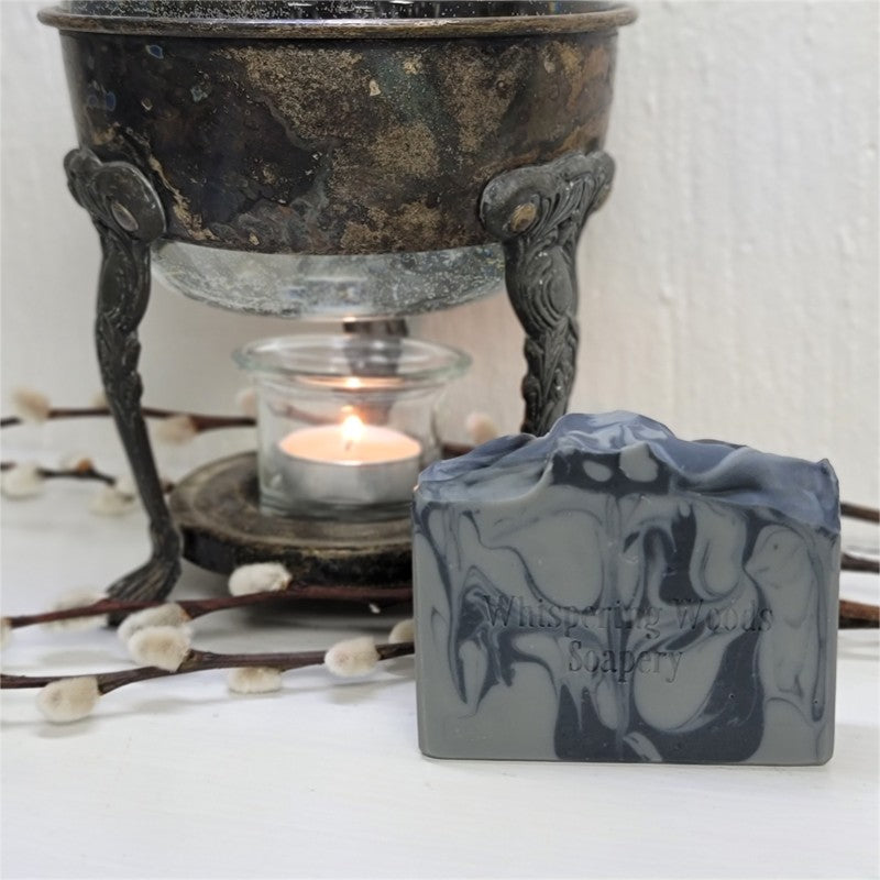 A rectangular bar of soap with a dark gray and black marbled swirl pattern sits in the foreground on a white surface. The soap is stamped with the brand name 'Whispering Woods Soapery.'