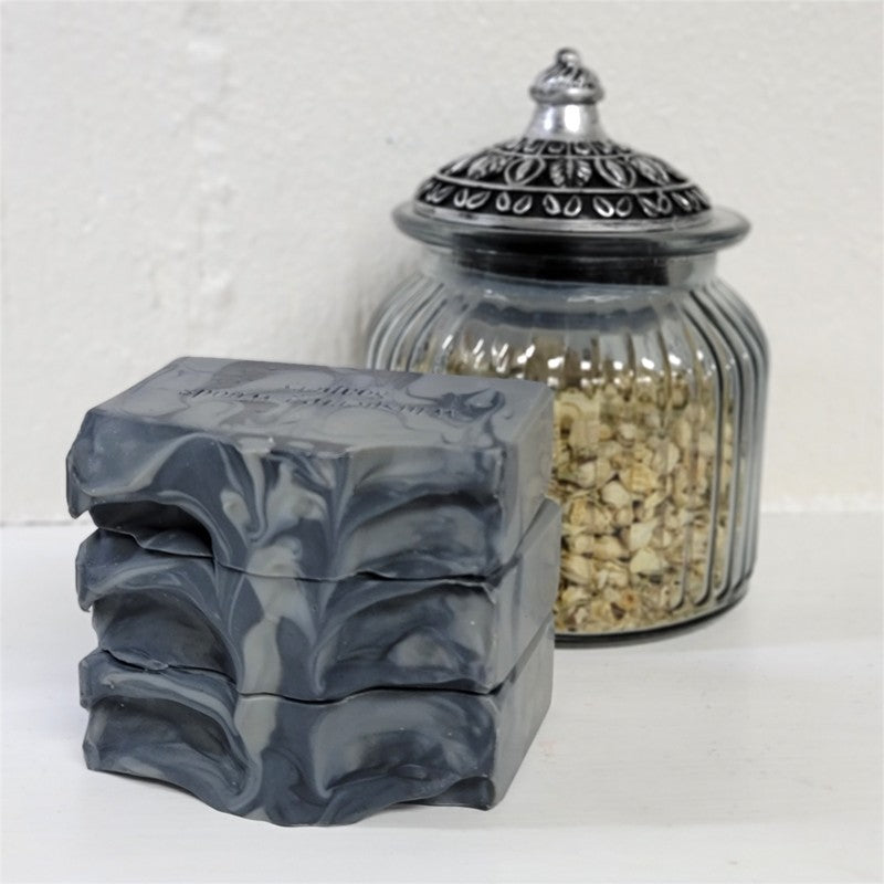 Three gray and black swirled bars of soap stacked on a white surface with a ribbed glass jar with a silver lid in the background.