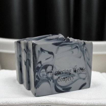Three bars of gray and black soap with 'Whispering Woods Soapery' branding on a white surface with a black curtain background