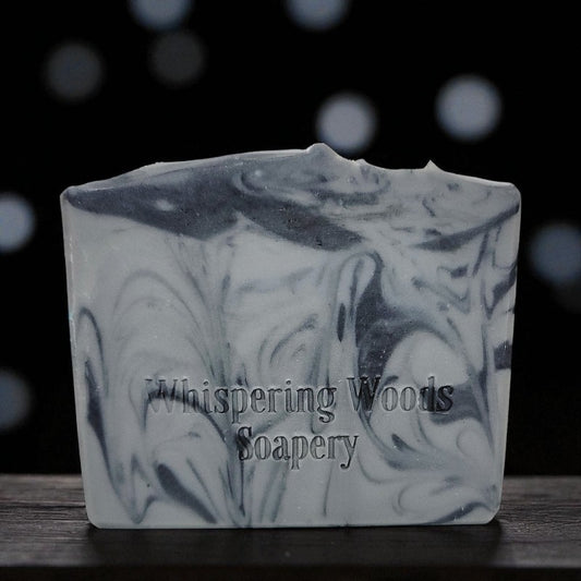 Bar of gray and black soap with swirling pattern and 'Whispering Woods Soapery' branding on a dark background