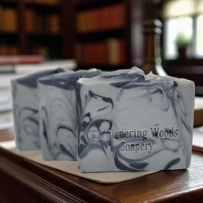 Three bars of soap with a marbled design on a wooden surface in a study, branded 'Whispering Woods Soapery.' 