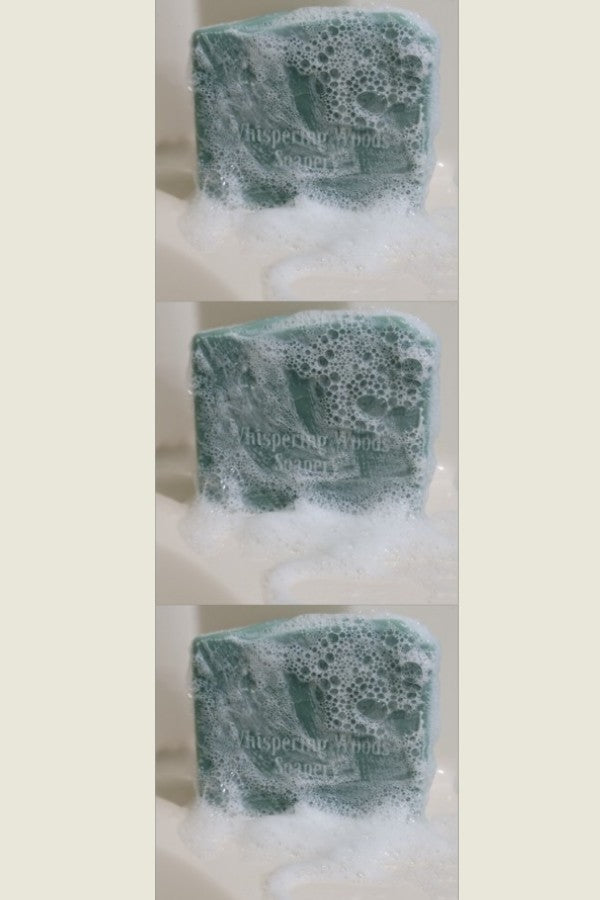 A vertical row of green soap set on the edge of a tub. They are stamped 'Whispering Woods Soapery' and are covered with bubbles and lather.
