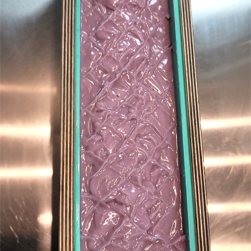 Close-up view of freshly poured purple soap batter in a rectangular mold. The textured top features a criss-cross hatch pattern. The mold sits on a stainless-steel surface.