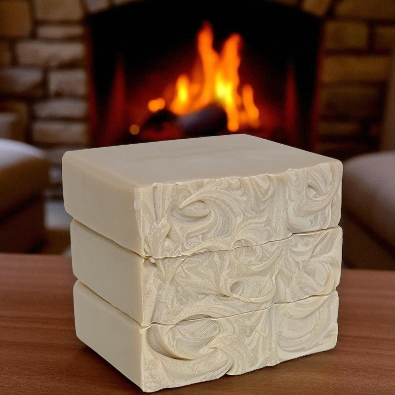Close-up of the swirled tops of a stack of light tan soap bars on a wooden surface with a fireplace in the background