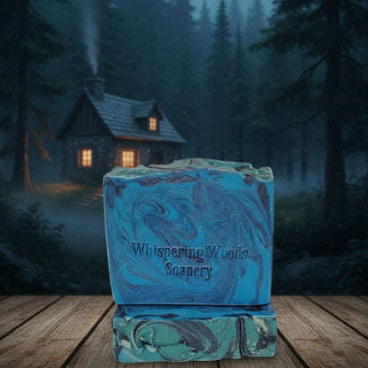 Blue soap bars with 'Whispering Woods Soapery' branding in front of a cottage in the woods