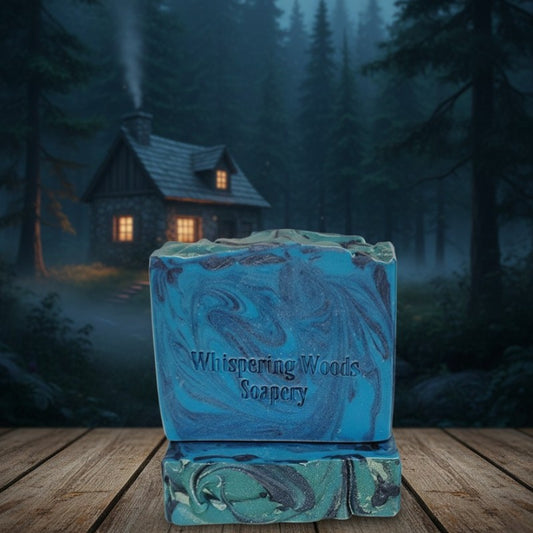 Blue soap bars with 'Whispering Woods Soapery' branding in front of a cottage in the woods
