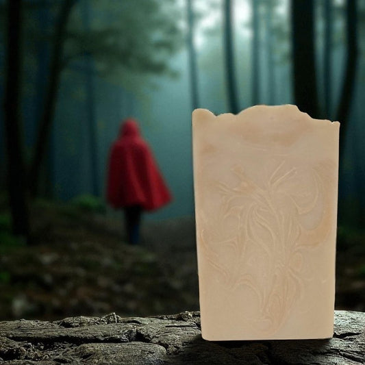 A red-hooded figure walks in the woods. There is a creamy tan soap with coppery swirls in the foreground.