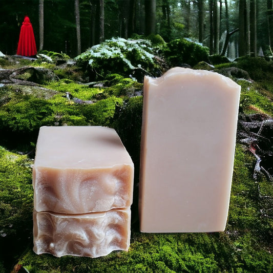 Three pale peach bars of soap on moss, with a forest in the background. A red cloaked figure can be seen amongst the trees in the background.